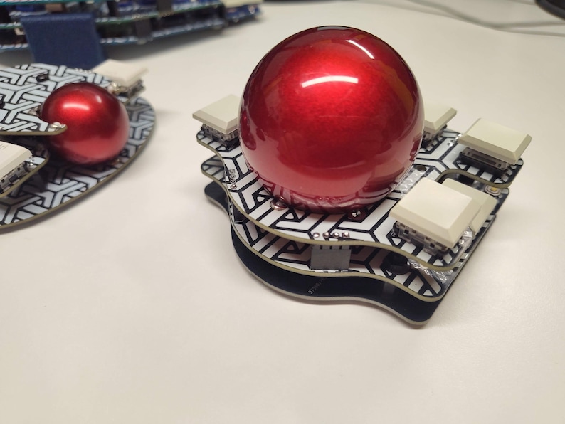 May include: A red, glossy sphere sits atop a layered, geometric patterned base with white square buttons. The base has a black border. Another similar object is visible in the background. The objects are on a white surface.