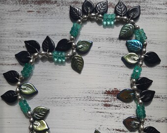 May include: A silver chain necklace with black and green glass leaf-shaped beads. The necklace is made of silver chain and has a clasp closure.