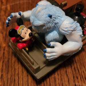 Puede incluir: Una figura de Mickey Mouse con un traje de esquí rojo en un trineo, perseguido por un Yeti azul claro. El Yeti tiene garras azules y está sobre una plataforma marrón con una maleta marrón.
