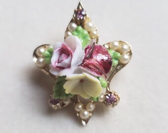 Puede incluir: Un broche de tono dorado con un grupo de flores de porcelana. Las flores son blancas, rosas y rojas con hojas verdes. El broche está decorado con pequeñas cuentas de perlas y diamantes de imitación rosas.