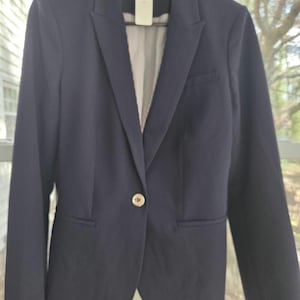 May include: A navy blue blazer with a single gold button closure. The blazer has a notched lapel and a pocket on each side. The interior lining features a striped pattern in red, white, and blue. The blazer is hanging on a black hanger.