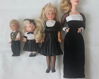 May include: Four dolls dressed in black and white formal attire. The dolls are standing on a white surface. The tallest doll has long blonde hair and is wearing a black velvet dress with a white sash. The other three dolls are wearing black dresses with white collars and bows. The smallest doll is a boy wearing a black suit with a patterned vest.