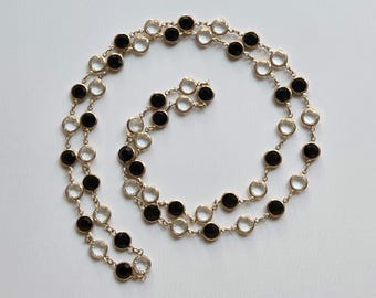 May include: A gold-toned necklace with alternating black and clear round gemstones. The necklace is laid out in a spiral pattern against a white background. The gemstones are evenly spaced and set in individual bezels.