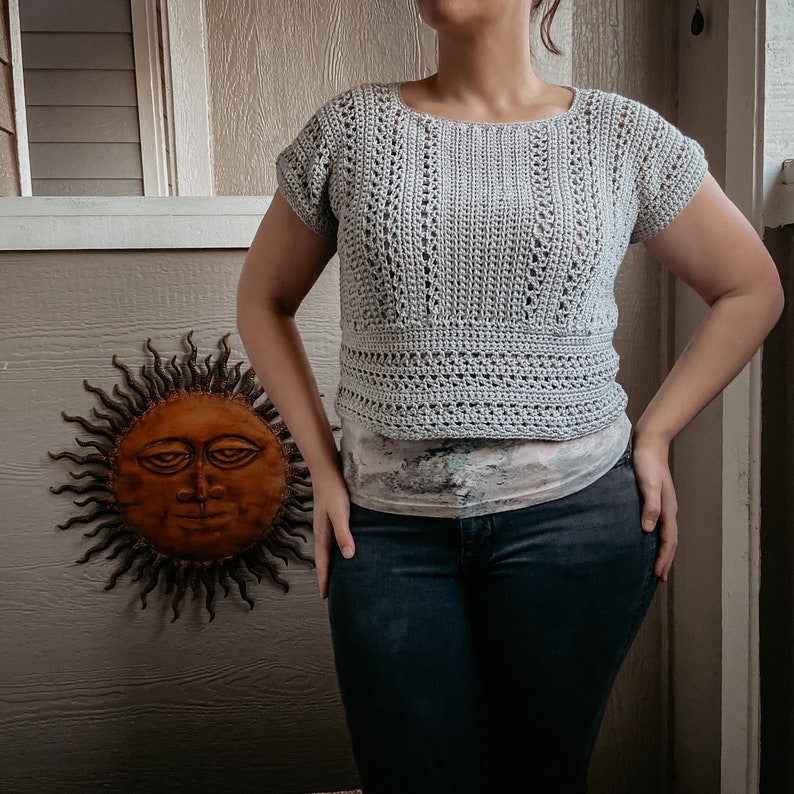 May include: A light gray crochet top with a loose, open-stitch pattern. The top has short sleeves and a cropped length. The model is wearing the top with a patterned undershirt and dark blue jeans.