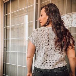 May include: A close-up of a woman wearing a light gray crochet top with a short sleeve. The top has a delicate openwork pattern and a slightly cropped length.