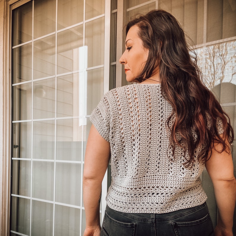 May include: A close-up of a woman wearing a light gray crochet top with a short sleeve. The top has a delicate openwork pattern and a slightly cropped length.