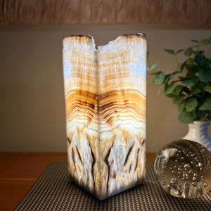 May include: A rectangular, illuminated onyx lamp with a natural, layered pattern of white, tan, and brown. The lamp is placed on a dark surface, next to a glass paperweight and a white vase with green foliage.