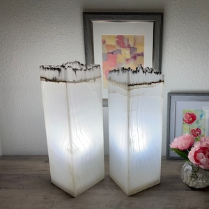 May include: Two white alabaster lamps with a black and white pattern on the top. The lamps are lit and casting a warm glow. The lamps are on a wooden surface with a vase of pink flowers and a framed picture in the background.