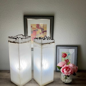May include: Two white, rectangular lamps with a wood-like texture and a black burnt edge. The lamps are lit and casting a warm glow. The lamps are on a wooden table with a framed painting and a vase of pink flowers.