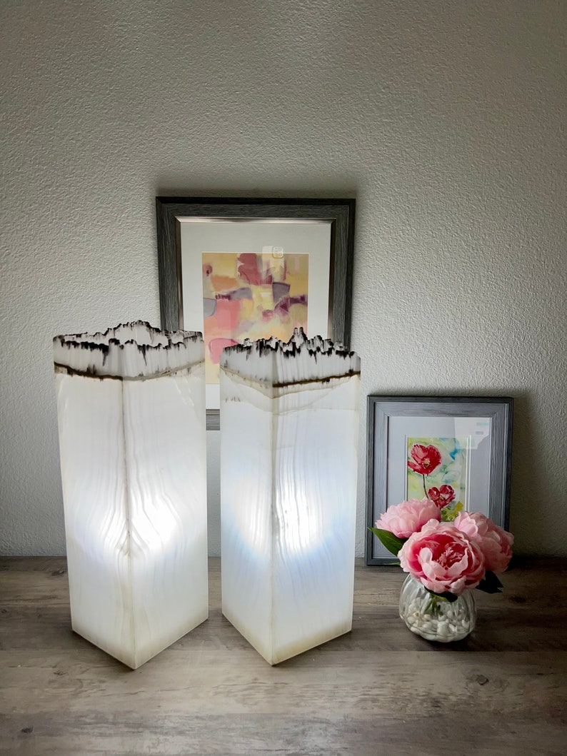 May include: Two white, rectangular lamps with a wood-like texture and a black burnt edge. The lamps are lit and casting a warm glow. The lamps are on a wooden table with a framed painting and a vase of pink flowers.
