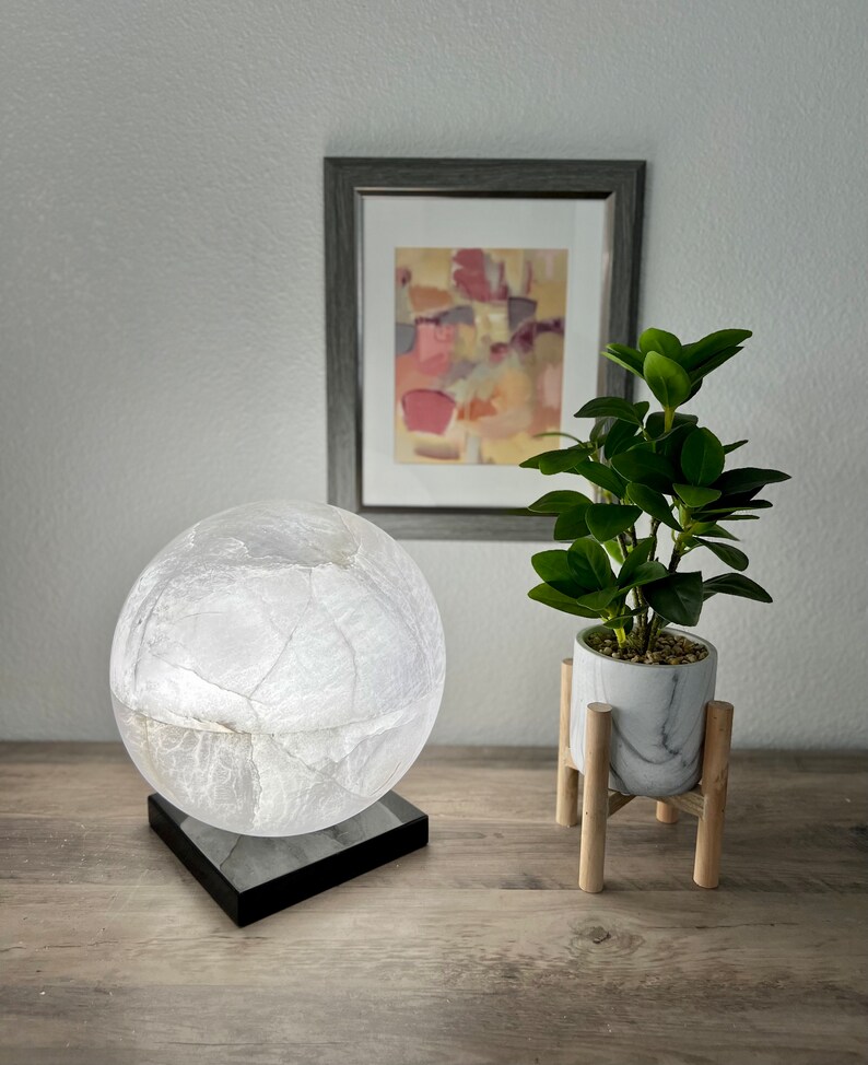 May include: A white globe lamp with a black base sits on a wooden table next to a potted plant on a wooden stand. The plant has green leaves and is in a white marble pot.