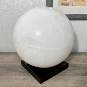 May include: A large white selenite sphere on a black base. The sphere has a smooth, polished surface with natural striations.