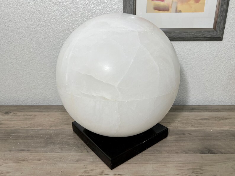 May include: A large white selenite sphere on a black base. The sphere has a smooth, polished surface with natural striations.