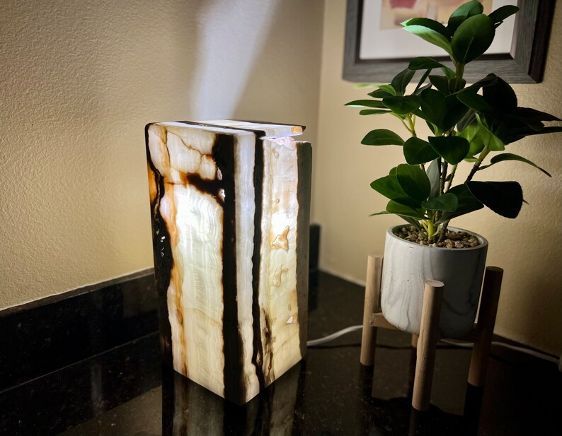 May include: A white and black onyx table lamp with a rectangular base. The lamp is lit and casting a warm glow.