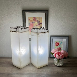 May include: Two white resin lamps with a wood grain pattern and a black resin top. The lamps are lit and casting a warm glow. The lamps are on a wooden table with a vase of pink flowers and two framed pictures in the background.