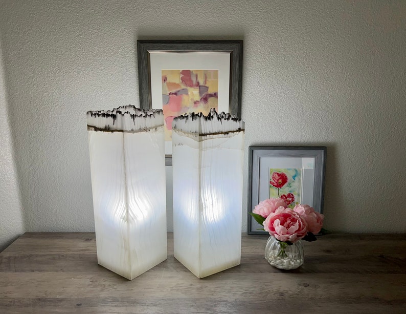 May include: Two white resin lamps with a wood grain pattern and a black resin top. The lamps are lit and casting a warm glow. The lamps are on a wooden table with a vase of pink flowers and two framed pictures in the background.