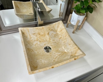 May include: A square, cream-colored stone vessel sink with a slightly curved edge. The sink has a subtle marbled pattern and a chrome drain.