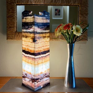 May include: A tall, rectangular lamp with a layered, multicolored design. The lamp features shades of blue, brown, orange, and white. A silver vase with flowers sits to the right of the lamp. A mirror with a woven frame hangs in the background.