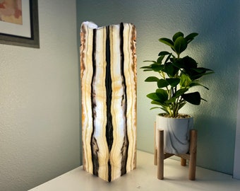 May include: A tall, rectangular onyx lamp with a natural, striped pattern in shades of brown, white, and black. The lamp is lit and casting a warm glow. It is sitting on a white surface next to a potted plant.