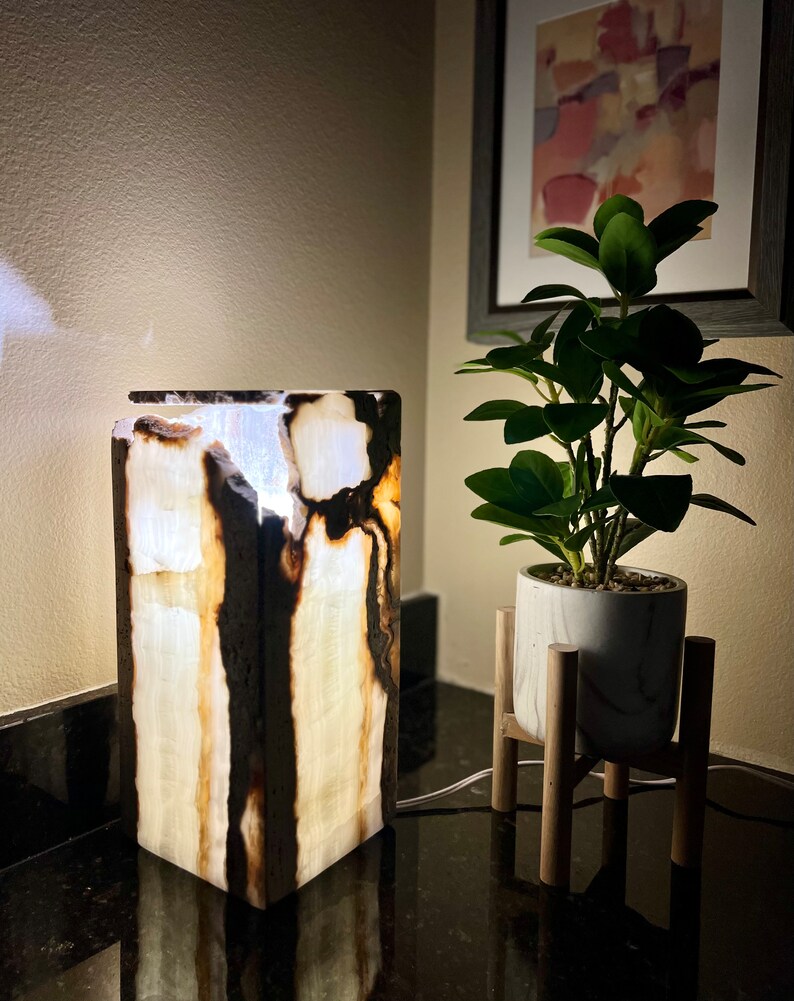 May include: A white and brown onyx lamp with a light inside. The lamp is on a black surface next to a potted plant on a wooden stand.