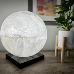 May include: A white, marble-like sphere lamp with a black base. The lamp is on a wooden table next to a potted plant.