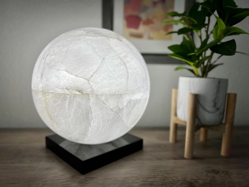 May include: A white, marble-like sphere lamp with a black base. The lamp is on a wooden table next to a potted plant.