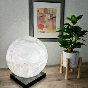 May include: A white alabaster sphere lamp with a black base. The lamp is on a wooden table next to a potted plant.