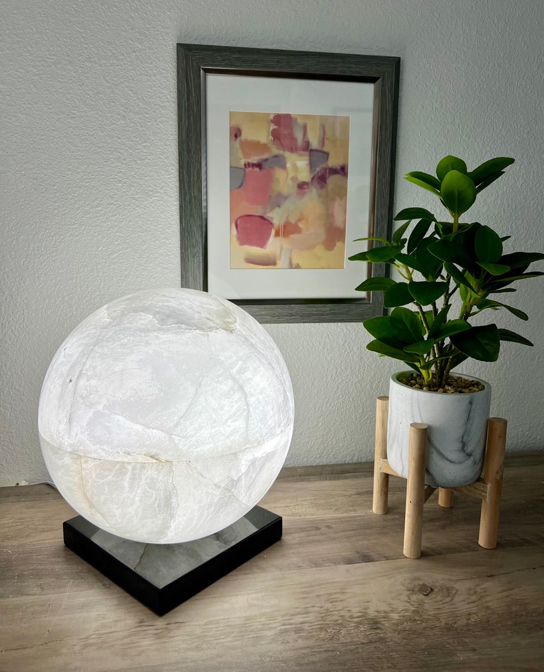May include: A white alabaster sphere lamp with a black base. The lamp is on a wooden table next to a potted plant.