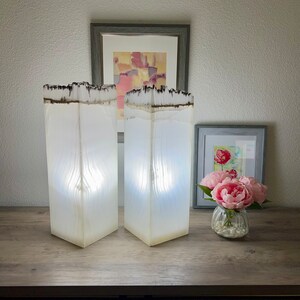 May include: Two white resin lamps with a wood grain texture and a dark brown top. The lamps are on a wooden surface with a picture frame and a vase of pink flowers.