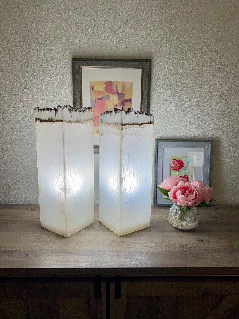 May include: Two white resin lamps with a wood grain texture and a dark brown top. The lamps are on a wooden surface with a picture frame and a vase of pink flowers.
