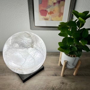 May include: A white moon lamp with a textured surface sits on a black base. The lamp is on a wooden table next to a potted plant with green leaves.