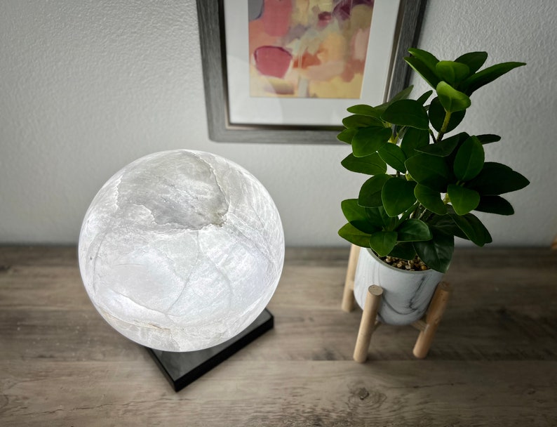 May include: A white moon lamp with a textured surface sits on a black base. The lamp is on a wooden table next to a potted plant with green leaves.