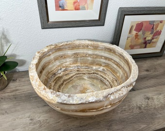 May include: A large, round, white and brown onyx bowl with a smooth, polished surface. The bowl has a natural, organic shape and is perfect for displaying decorative items or serving snacks.