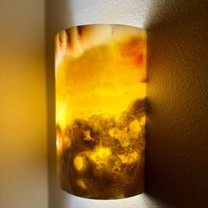 May include: A semi-cylindrical wall sconce crafted from translucent stone, featuring warm amber and golden hues with natural veining patterns. The light source illuminates the stone from within, creating a soft glow that highlights the organic textures and layered mineral formations across the curved surface.