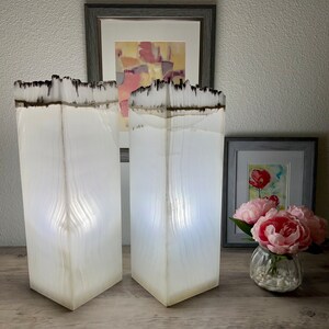 May include: Two white alabaster lamps with a wood grain pattern and a black top. The lamps are lit and casting a warm glow. The lamps are on a wooden surface.