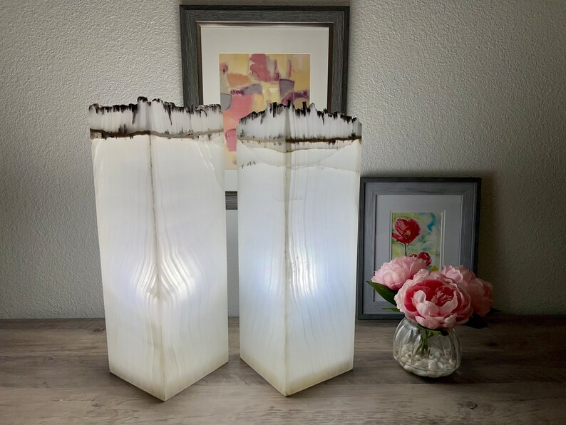 May include: Two white alabaster lamps with a wood grain pattern and a black top. The lamps are lit and casting a warm glow. The lamps are on a wooden surface.