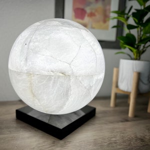 May include: A white, spherical lamp with a textured surface sits on a black square base. The lamp is illuminated, casting a soft glow.