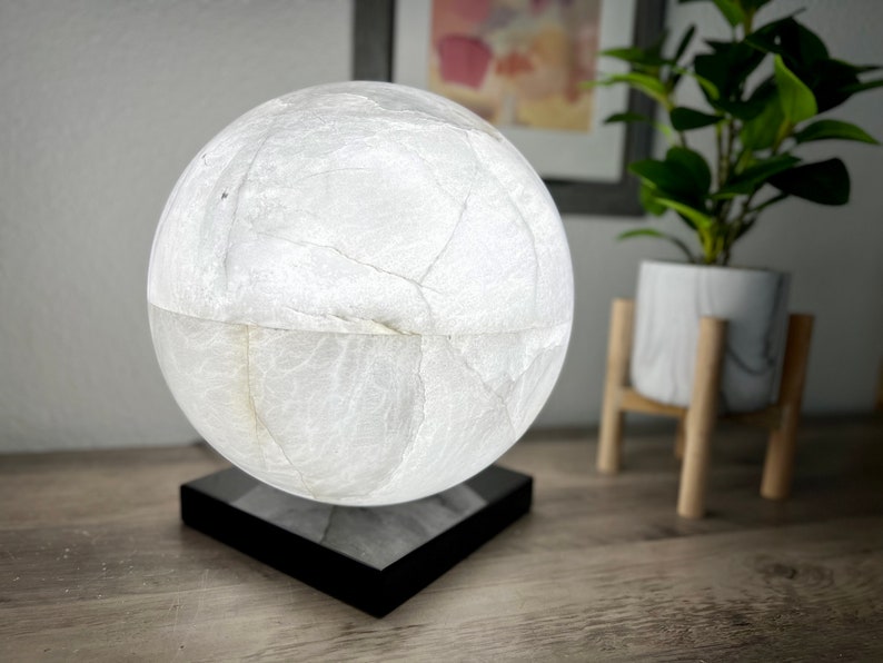 May include: A white, spherical lamp with a textured surface sits on a black square base. The lamp is illuminated, casting a soft glow.