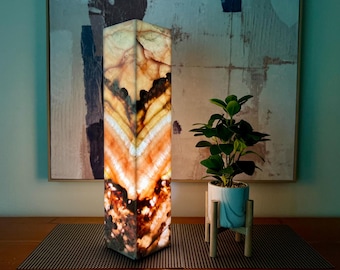 May include: A tall, rectangular, onyx lamp with a warm, amber glow. The lamp is made of natural stone with brown, orange, and white patterns. It is sitting on a wooden table with a brown placemat.