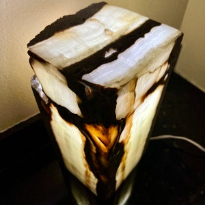 May include: A white and black onyx lamp with a unique, natural, and organic design. The lamp is lit and the light shines through the onyx, creating a warm glow.