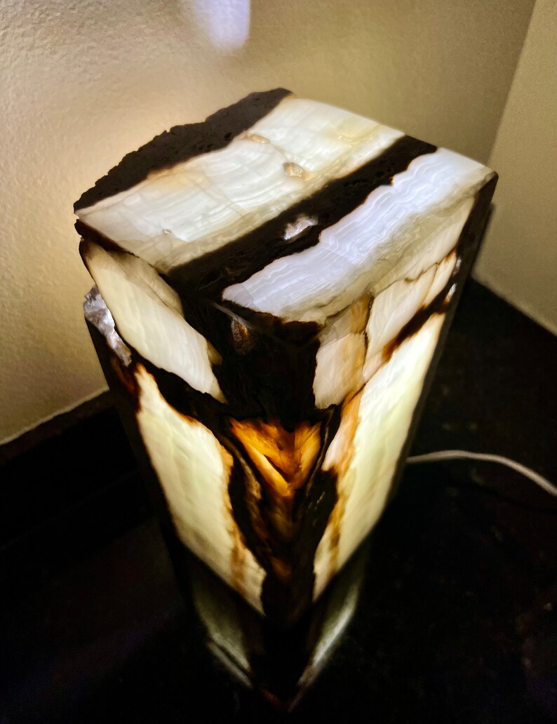 May include: A white and black onyx lamp with a unique, natural, and organic design. The lamp is lit and the light shines through the onyx, creating a warm glow.