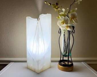 May include: A white, translucent, sculptural lamp with a modern design sits on a white table next to a vase with white flowers. The vase is made of clear glass and has a black metal stand.