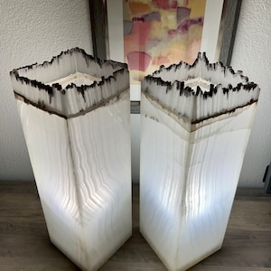 May include: Two white alabaster lamps with a black burnt edge. The lamps are square and have a textured surface. The lamps are lit and casting a warm glow.