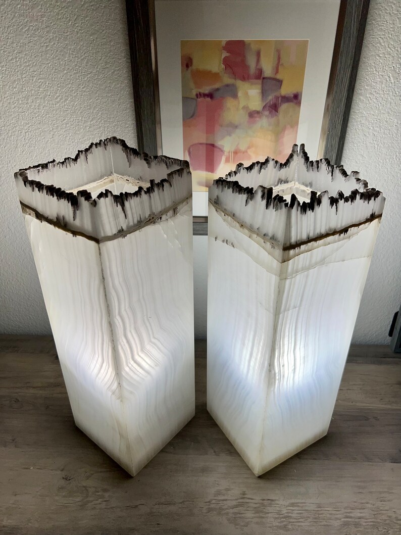 May include: Two white alabaster lamps with a black burnt edge. The lamps are square and have a textured surface. The lamps are lit and casting a warm glow.