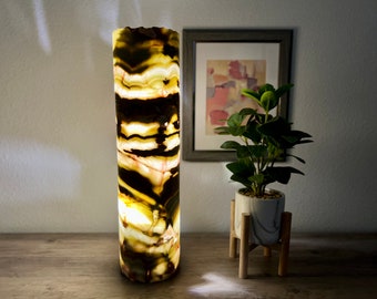 May include: A tall, cylindrical lamp made of onyx with a swirling pattern of brown, green, and white. The lamp is lit and casting a warm glow. The lamp is on a wooden surface.
