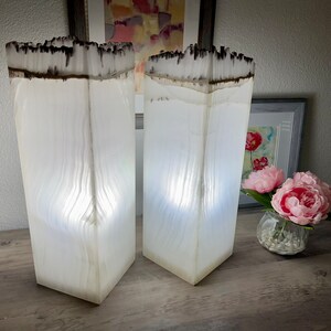 May include: Two white, rectangular lamps with a wood grain pattern and a black top. The lamps are lit and casting a warm glow.