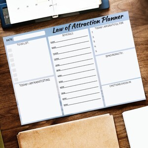 May include: A white planner with a blue header that says "Law of Attraction Planner". The planner is divided into sections for scheduling, to-do list, today I am manifesting, today I am grateful for, synchronicity, and emotions check-in.