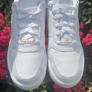 May include: A pair of white sneakers featuring gold-toned metal lace charms. The left shoe displays the name Sarah in a cursive script, while the right shoe displays the name Alex in a matching cursive script, both threaded onto the white laces.