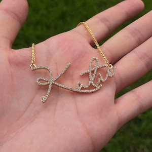 May include: A gold necklace with the name "Katie" spelled out in cursive script with small clear rhinestones.