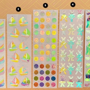 Pode incluir: Dez folhas de adesivos holográficos coloridos com vários desenhos. Os adesivos incluem cisnes, veleiros, carinhas sorridentes, coelhos com tutu e uma cena de natividade.